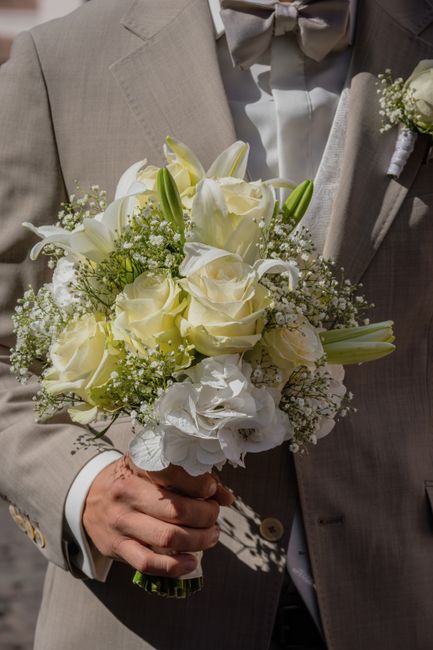 A close-up view of a bride or groom holding a bouquet arrangement of predominantly white flowers, including roses, lilies, and hydrangeas, accented with small white baby's breath and green foliage. Th
