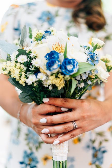A bride holds a bouquet arrangement featuring white and blue flowers, including roses, delphiniums, and daisies, accented with green foliage. The bouquet is wrapped with white ribbon, and the bride’