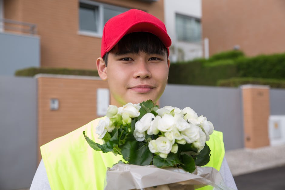 A young delivery person wearing a red cap and a high-visibility yellow vest is holding a bouquet of white roses wrapped in white paper. The flowers display fresh, tightly closed petals and glossy gree