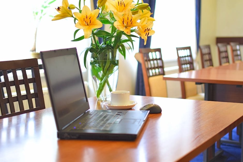 An office table featuring a clear glass vase filled with a floral arrangement of vibrant yellow lilies and green foliage, positioned centrally. The arrangement is fresh, with open petals and sturdy st