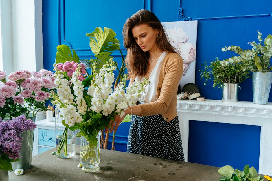 A female florist arranging a large bouquet of fresh white snapdragons, pink chrysanthemums, and greenery in a clear glass vase inside a bright, well-lit flower shop with vibrant blue walls. The arrang